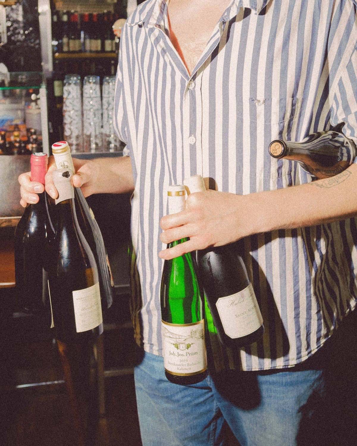 Fyra vinflaskor burna i famnen framför vinhyllan i baren.